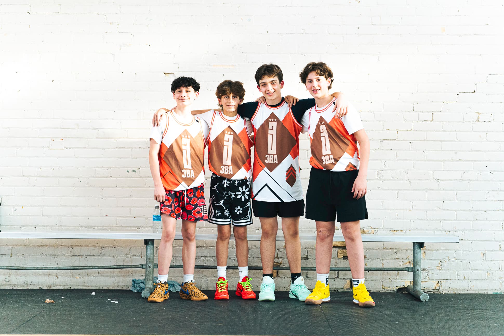 Four teammates in orange and white 3BA jerseys with their arms around each other against a white brick wall